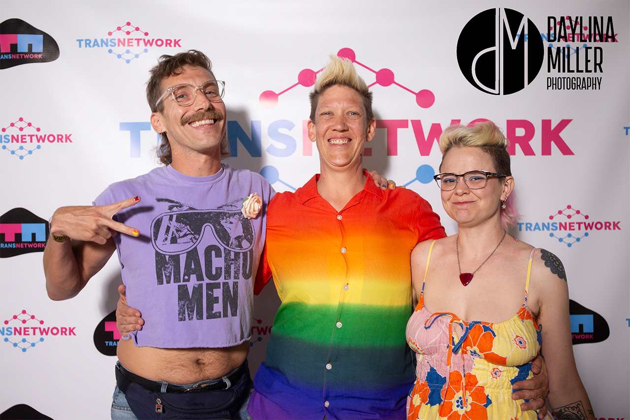 Adam and friends at the UNI-Tea Dance, smiling and posing against a TransNetwork backdrop